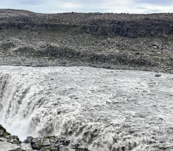 A vast, powerful waterfall cascades over a rocky cliff under a cloudy sky in one of the most Instagrammable places in Iceland, surrounded by a rugged landscape.