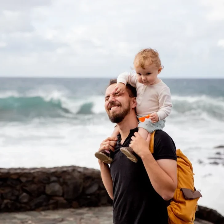 A joyful father traveling with his toddler, carrying him on his shoulders by the sea, both smiling, with waves crashing in the background.