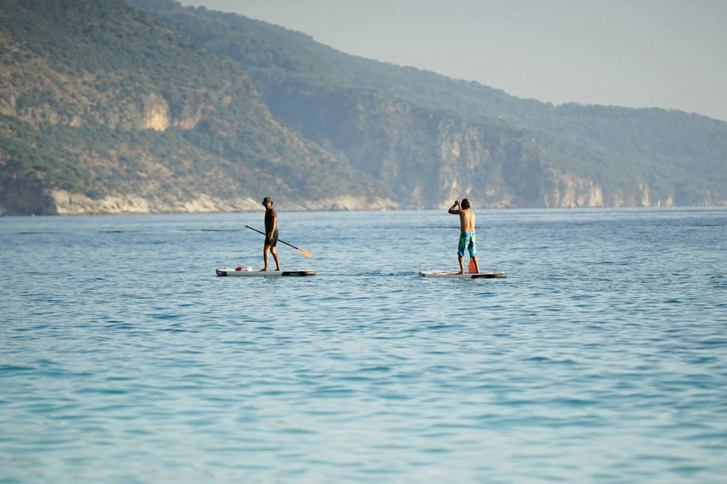 Two people paddleboarding on calm sea near hills