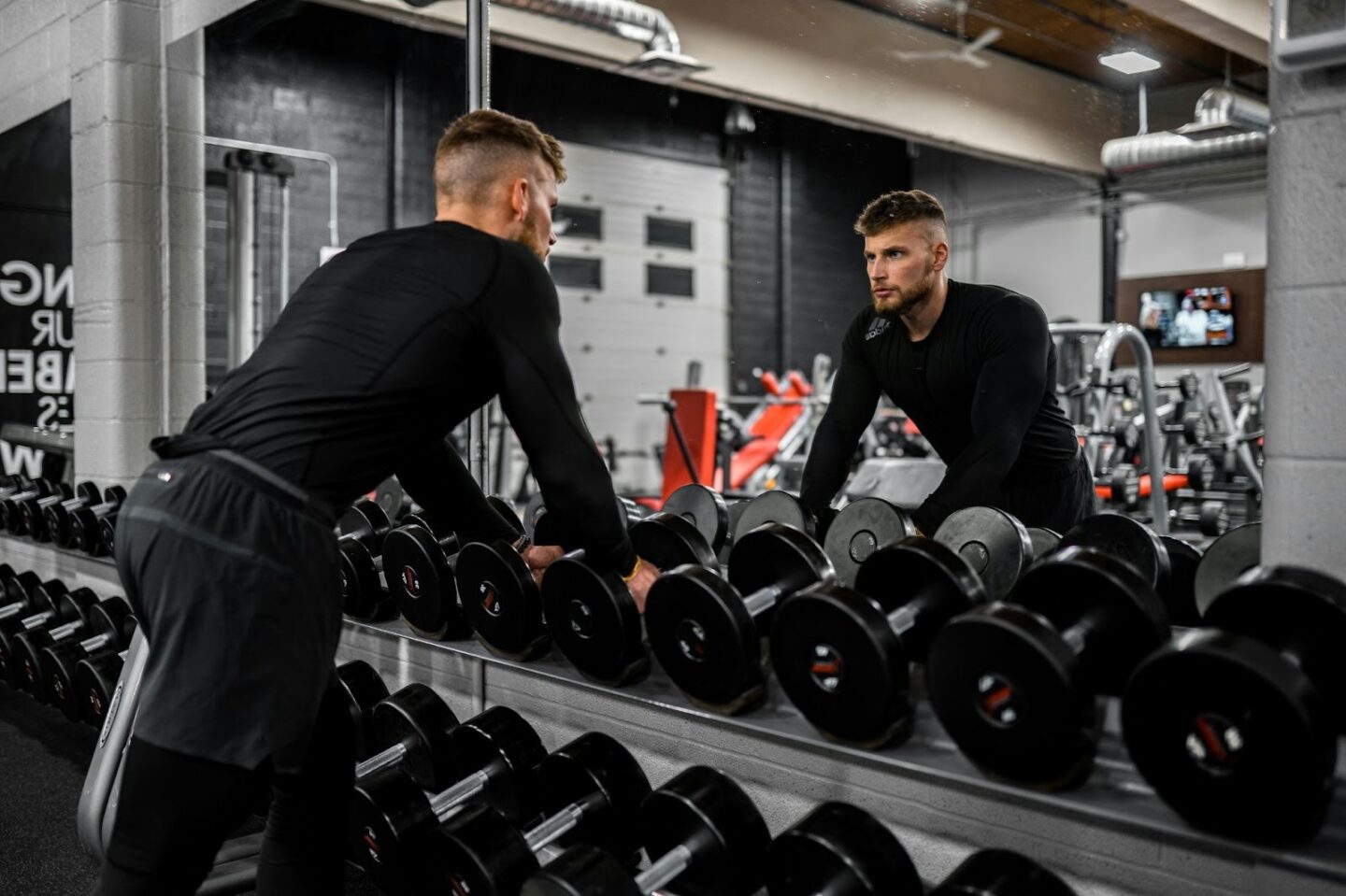 Man selecting dumbbells in gym, reflection in mirror